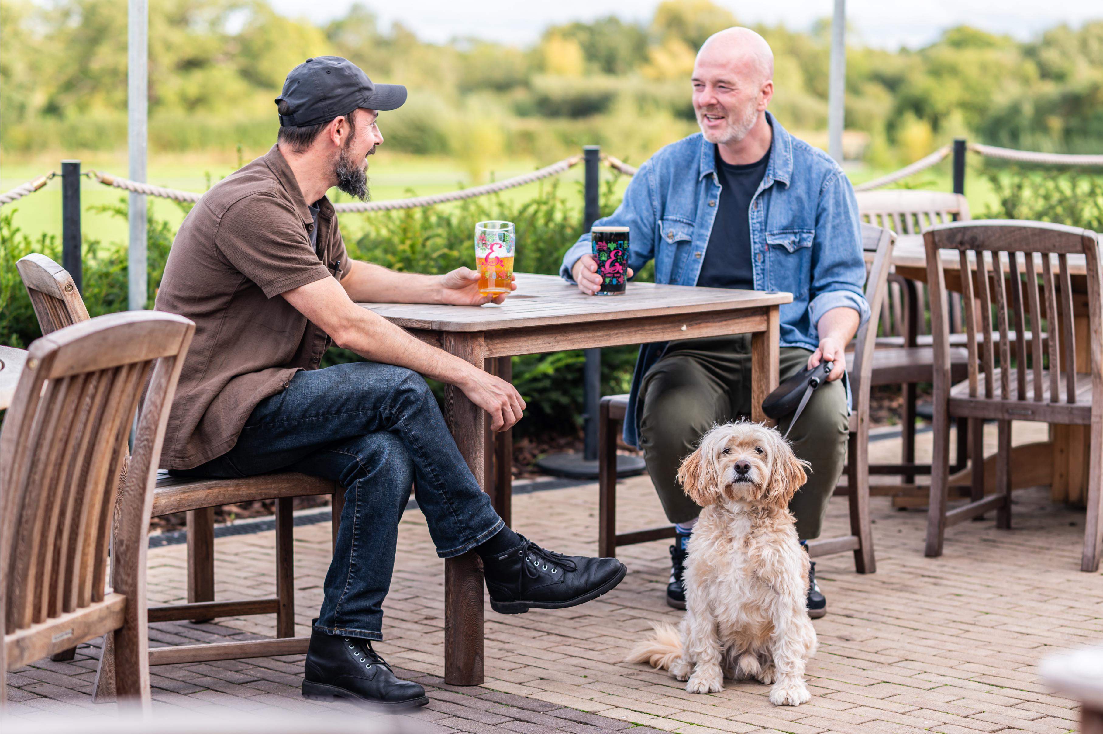 two men having a drink at a beer garden with thier dog